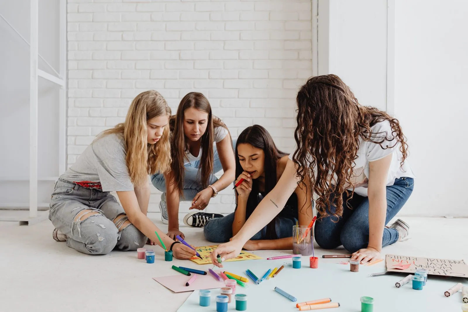 Four teenage girls collaboratively painting and drawing on a floor with bright colors, embracing creativity.